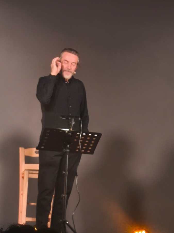 Un uomo in abbigliamento scuro parla davanti a un leggio, esibendosi in un contesto teatrale.