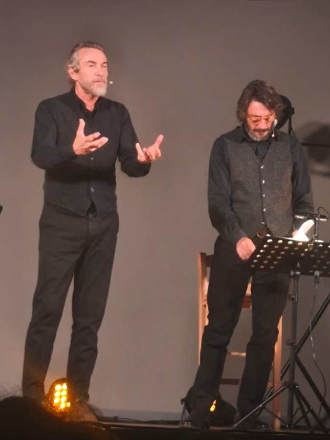 Due attori sul palco durante una performance teatrale, con un'illuminazione calda e un'atmosfera coinvolgente.