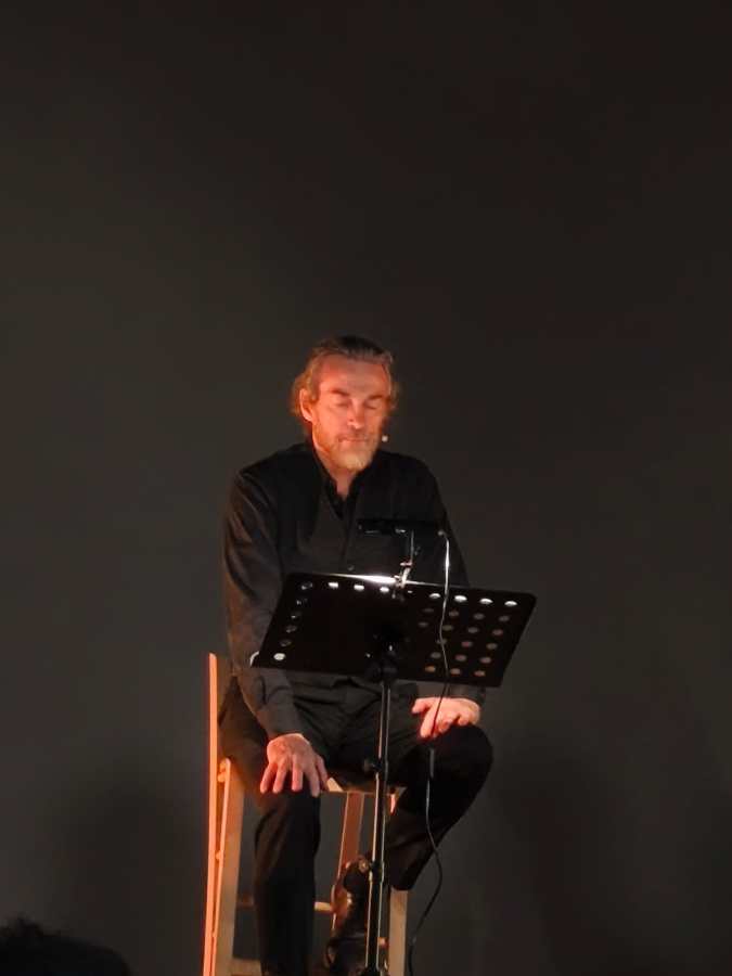 Un attore seduto su uno sgabello, mentre legge da un leggio in un teatro.