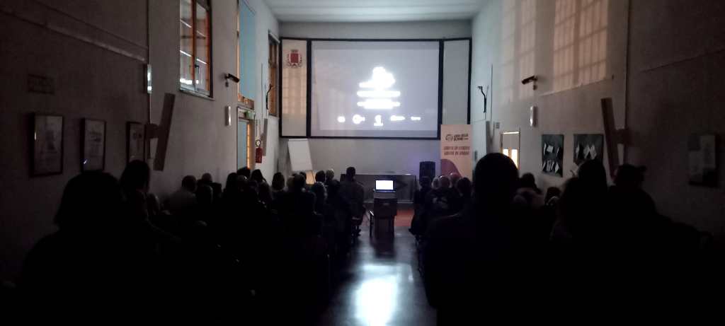 Interno di una sala con un pubblico seduto mentre assiste a una proiezione su uno schermo grande.