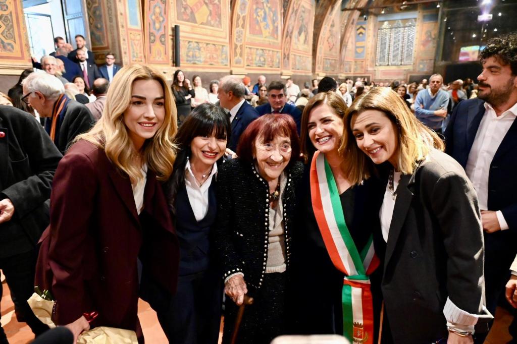 Gruppo di donne sorridenti, tra cui Mirella Alloisio, mentre festeggiano la sua cittadinanza onoraria a Perugia, all'interno della storica Sala dei Notari. La scena è vivace e affollata, con un pubblico che assiste alla cerimonia.