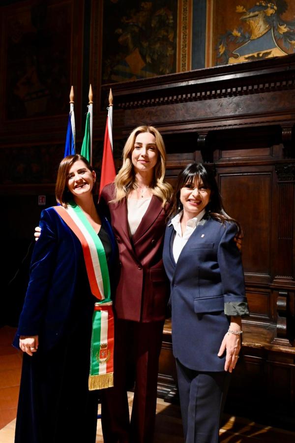 Tre donne sorridenti posano insieme durante una cerimonia ufficiale, con bandiere italiane sullo sfondo. Una di loro indossa una giacca blu scuro e una fascia tricolore, probabilmente una sindaca, mentre le altre sono in abbigliamento formale.