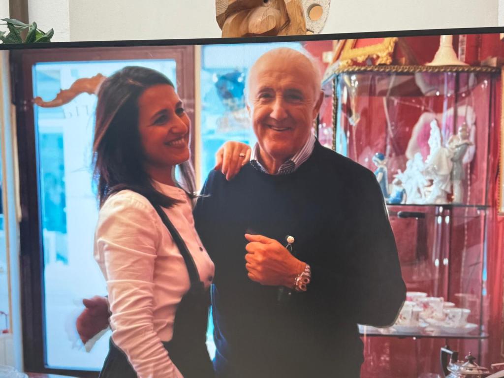 A smiling woman and man pose together in an antique shop, surrounded by decorative items in display cases.