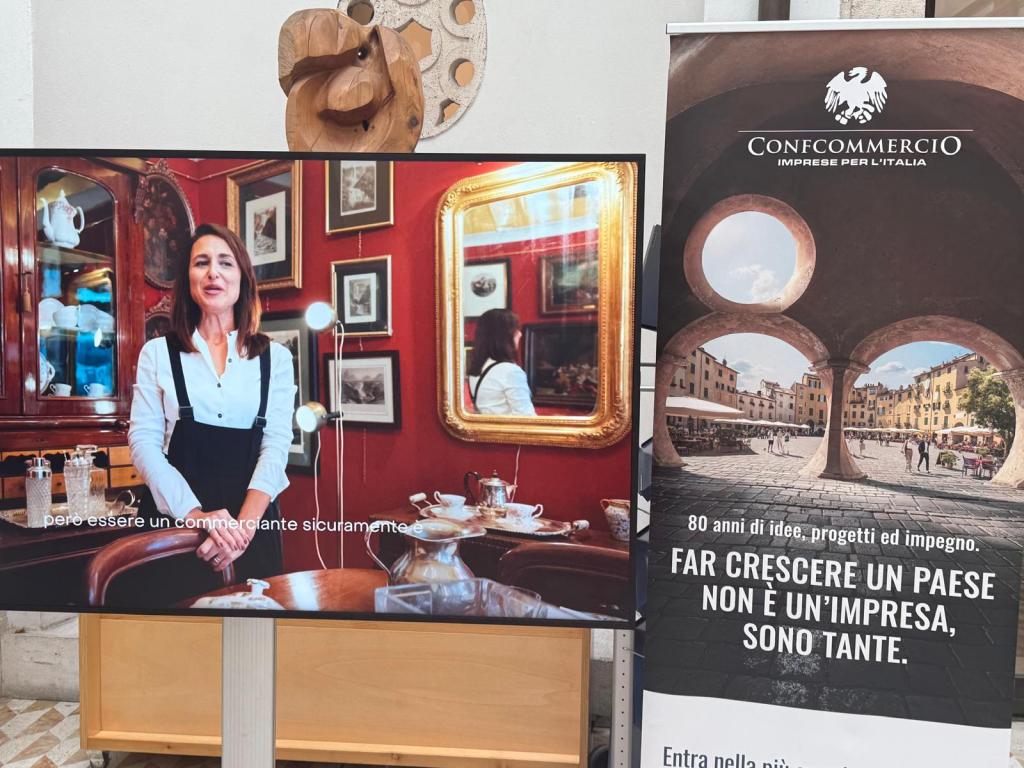 A woman stands in front of a decorative wooden cabinet filled with glassware and framed art, smiling while wearing a white shirt and black suspenders. Next to her is a promotional banner for Confcommercio, displaying a scenic image of an Italian town and the text about business and community growth.