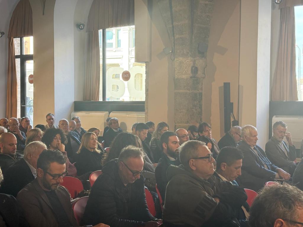 A crowded auditorium with people sitting in chairs, engaging in a meeting or presentation. Natural light coming through windows illuminates the attendees.