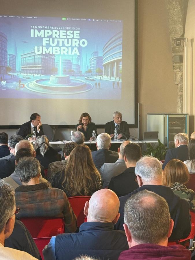 A conference setting with three speakers at a table, discussing the event titled 'Imprese Futuro Umbria' projected in the background. The audience is seated and engaged.