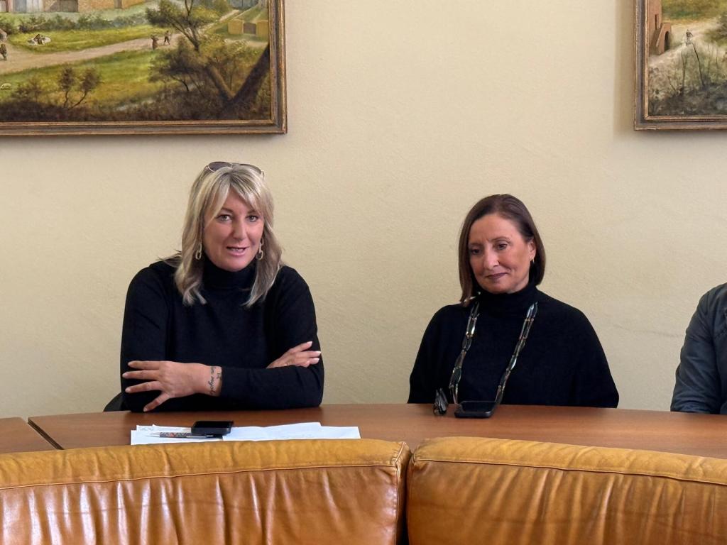 Two women seated at a table, both wearing black outfits. One woman has long blonde hair and earrings, while the other has medium-length brown hair. A painting is visible in the background.