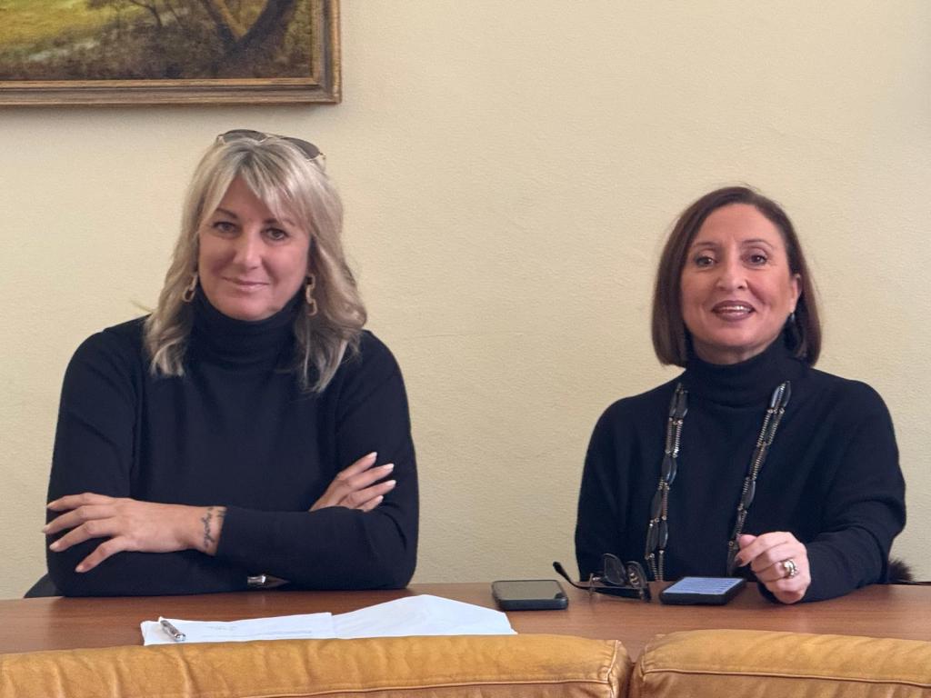 Two women seated at a table, both wearing black turtlenecks, with one woman smiling and the other resting her arms crossed. A notepad and a smartphone are visible on the table.