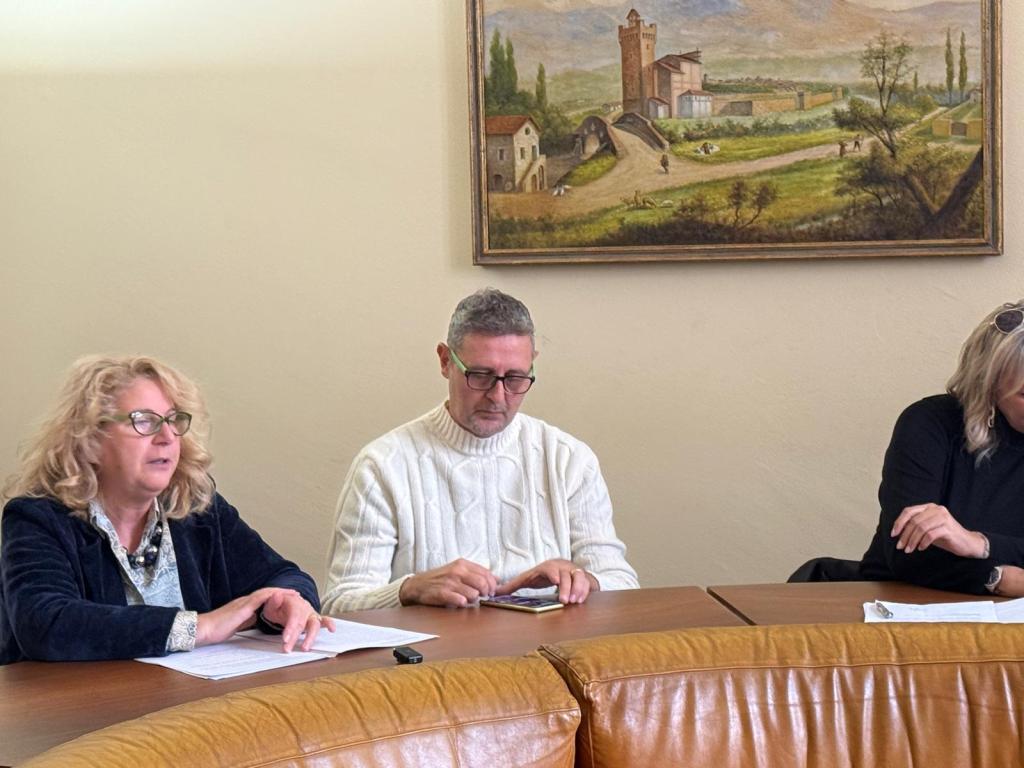 A group of three individuals sitting around a circular table, with one person speaking. There is a scenic painting of a landscape in the background.