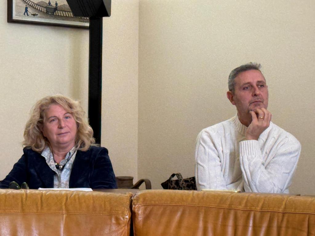 Two individuals seated on a couch, one with curly hair in a dark jacket and the other with short hair in a white sweater, both appear to be engaged in thought.