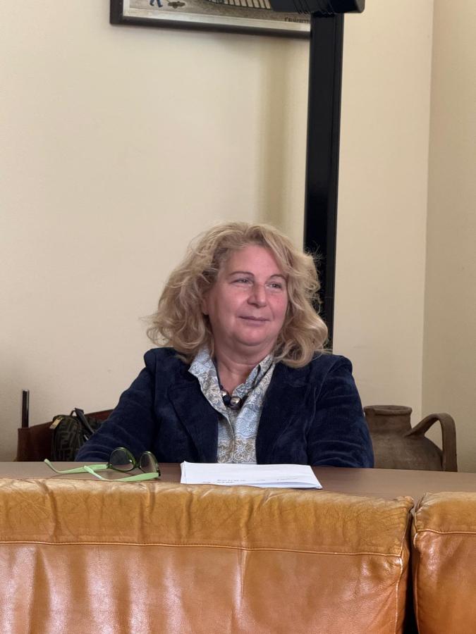 A woman with wavy blonde hair wearing a blue jacket, seated at a table with papers in front of her and green sunglasses.