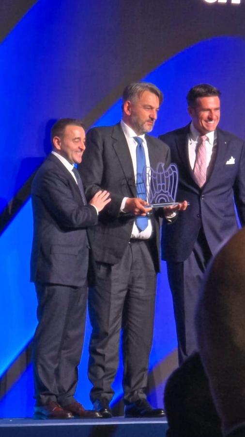 Tre uomini sorridenti sul palco, due in abito scuro e uno in camicia bianca con cravatta, mentre uno di loro tiene un premio in mano.