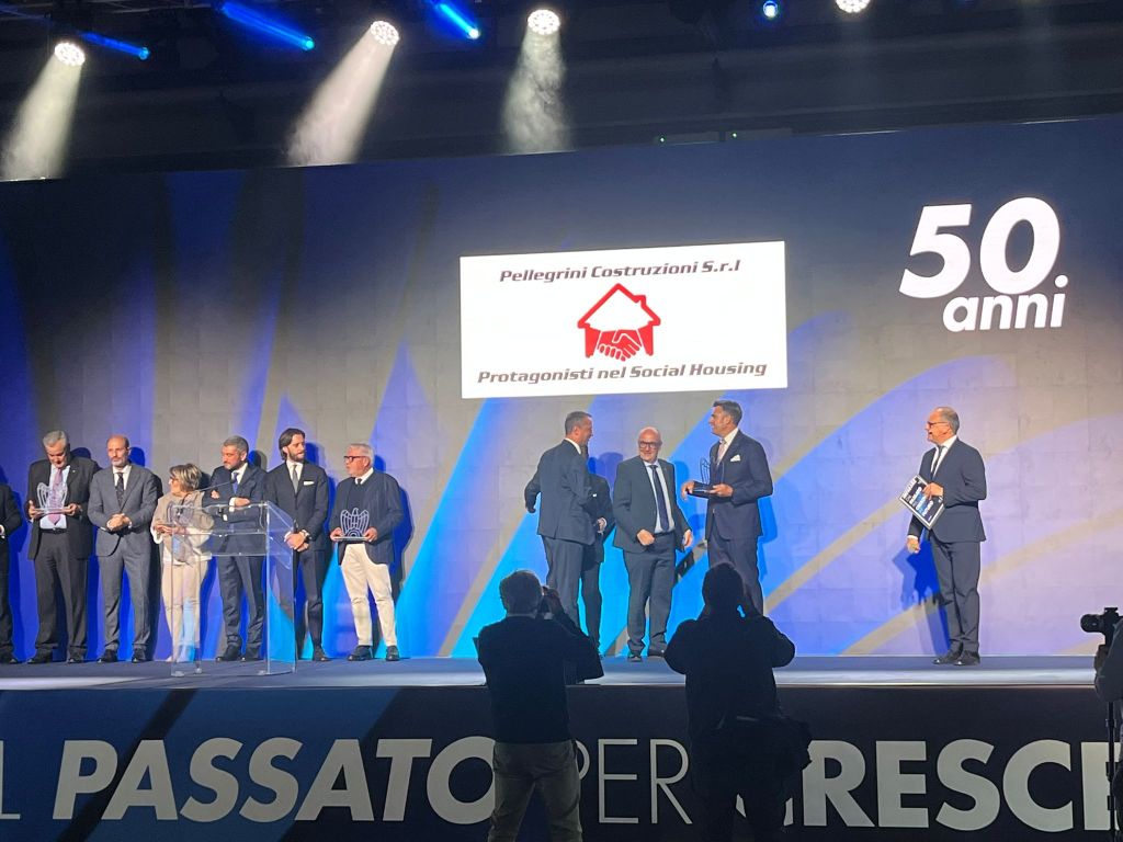 Cerimonia di premiazione con diverse persone sul palco che celebrano il 50° anniversario dell'impresa Pellegrini Costruzioni S.r.l., con un logo che rappresenta il Social Housing sullo sfondo.
