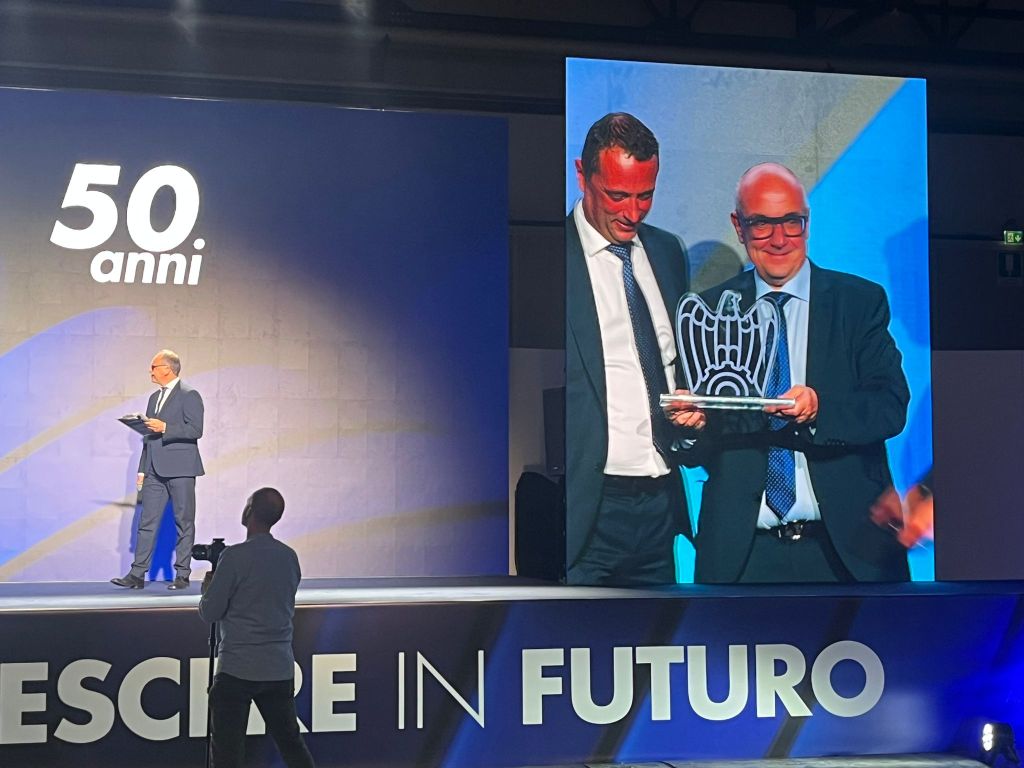 Cerimonia di premiazione per le imprese associate a Confindustria, con un relatore sul palco e un premiato che riceve un riconoscimento, durante un evento celebrativo.