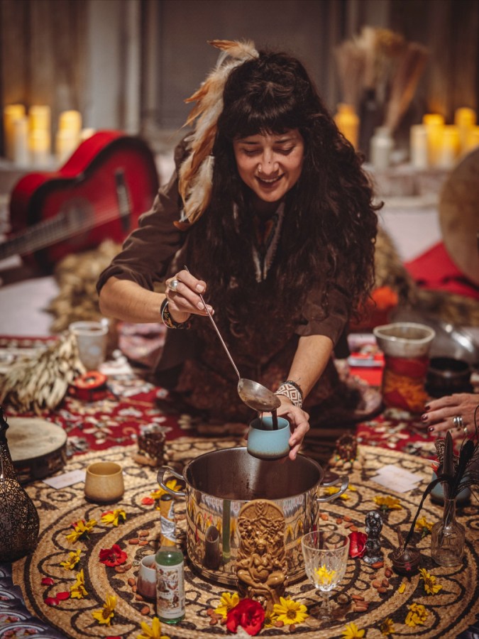 Una persona che mescola una bevanda in un pentolone, circondata da decorazioni artistiche e fiori, in un ambiente intimo e riflessivo.