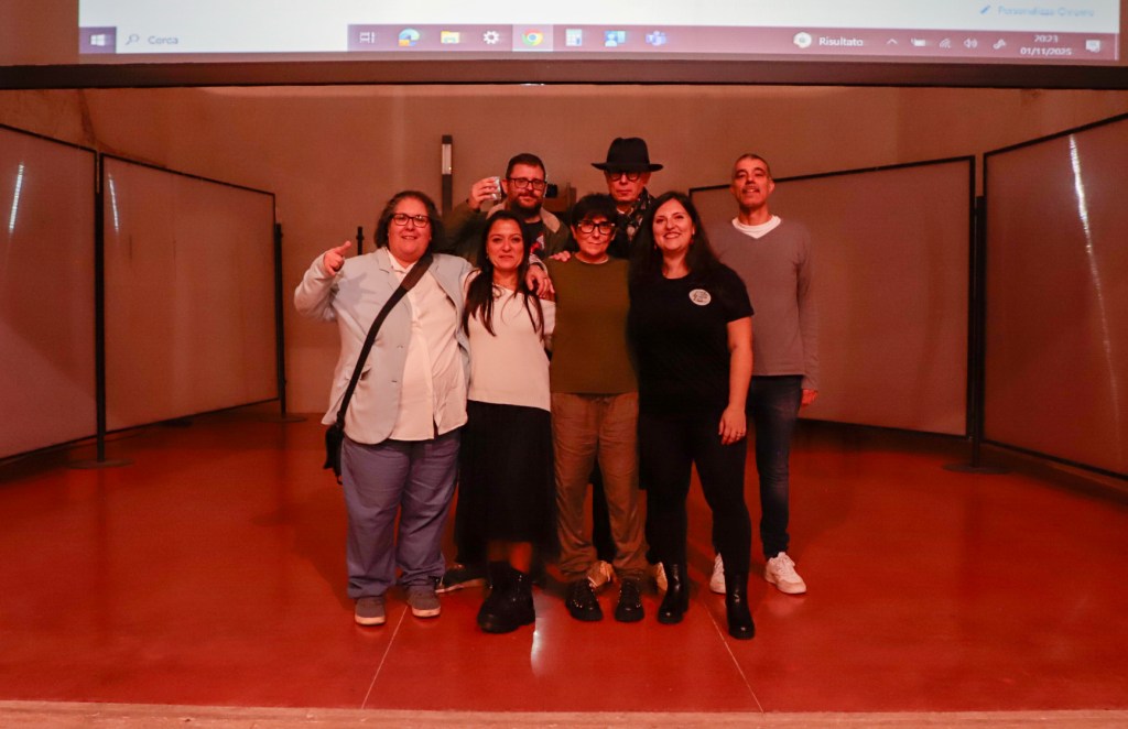 Un gruppo di otto persone in posa sorridente su un palco, in un evento legato al Terni e Narni Horror Fest, con uno sfondo di pannelli bianchi e uno schermo proiettore.