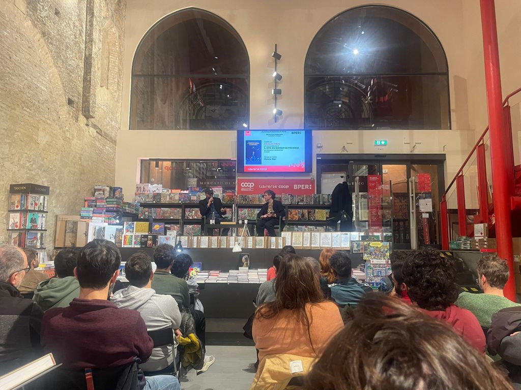 A bookshop event featuring two speakers on stage, with an audience seated in front. The interior has high arching windows and shelves filled with books.