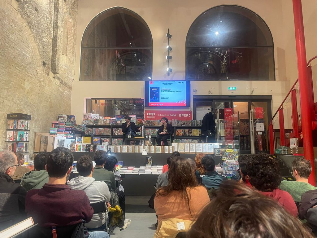 An audience seated in a bookstore attending an event, with two speakers on a stage in front of shelves filled with books.