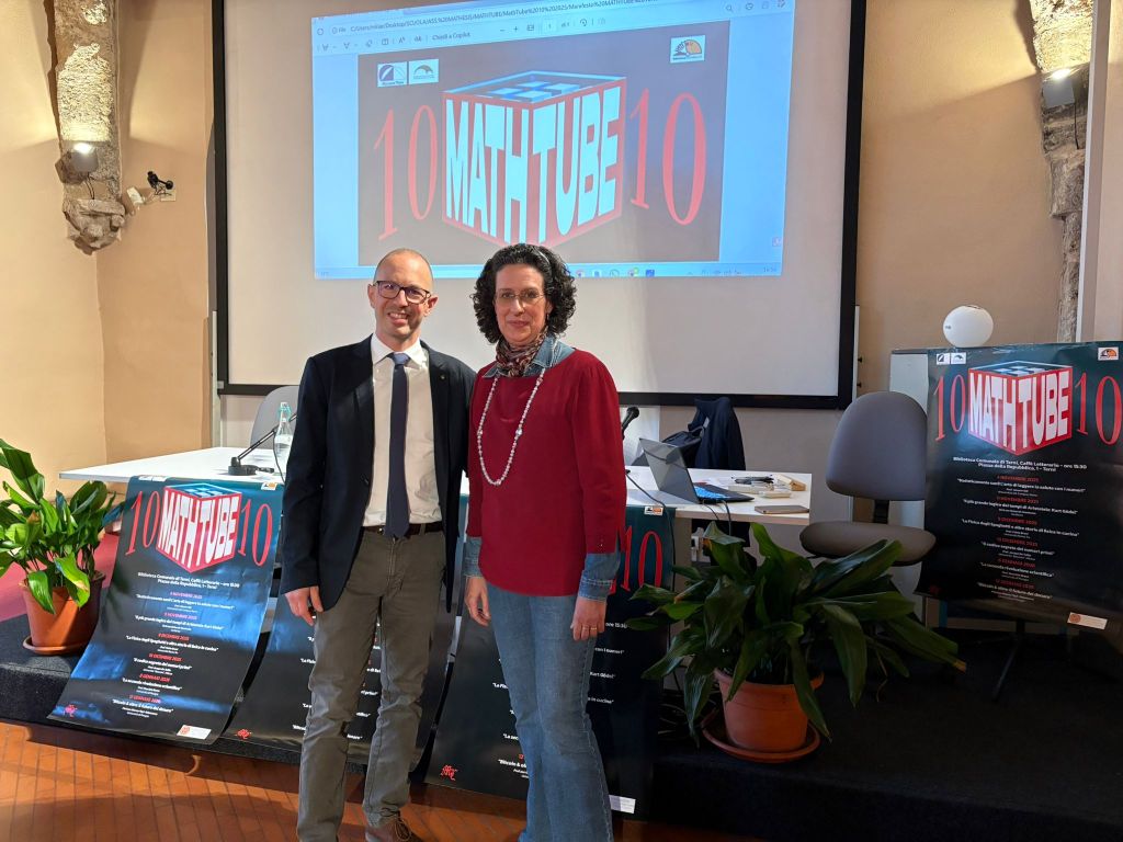 Due relatori sorridenti in piedi davanti a un podio, con un grande schermo e poster che pubblicizzano l'evento 'MathTube 10' sullo sfondo.