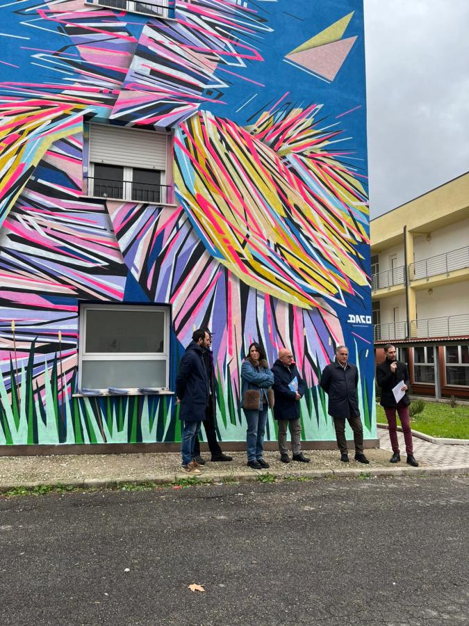 Inaugurazione del murales "GRAFFAUNE AMOUR" realizzato dall'artista DACO, che raffigura colori vivaci e geometrie, mentre un gruppo di persone osserva l'opera.