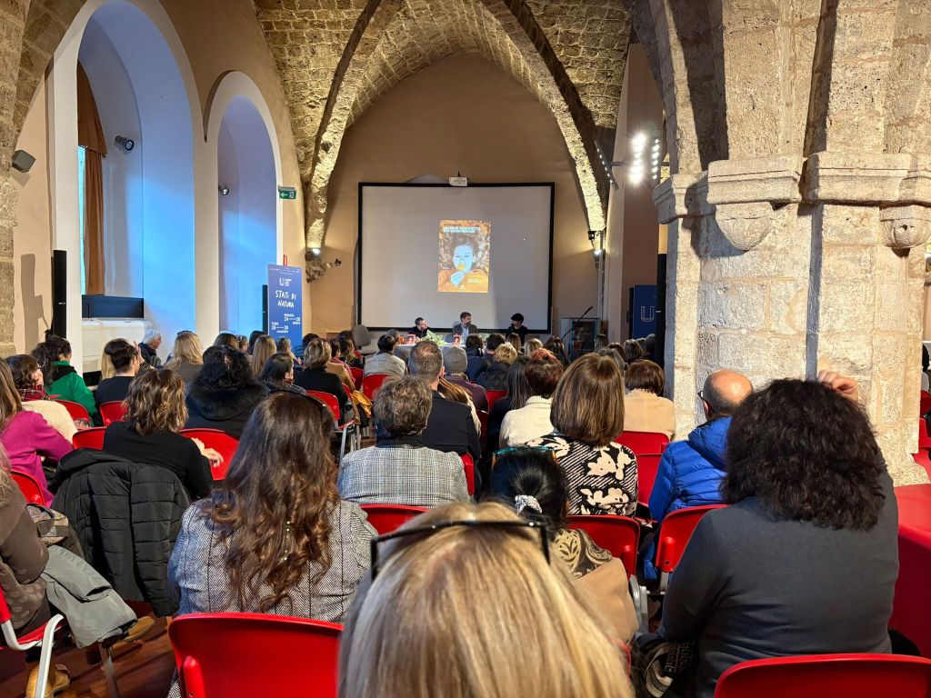 A crowded event space with an audience facing a stage where two speakers are discussing in front of a large projector screen displaying a book cover.