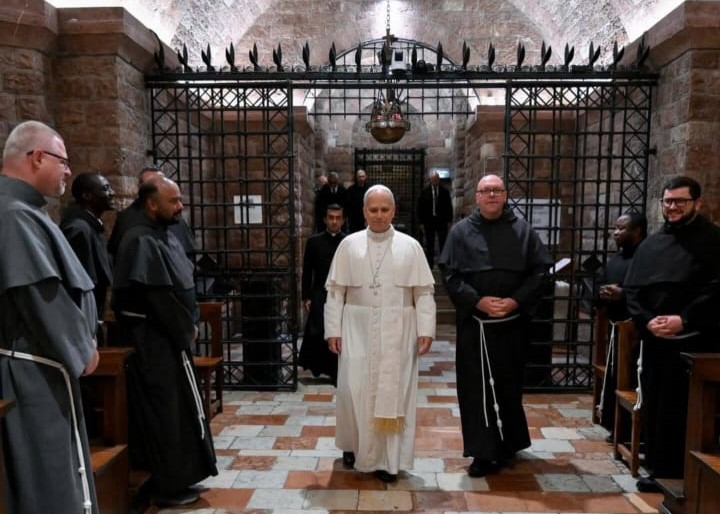 Papa Leone XIV in abiti papali cammina all'interno di un luogo sacro, accompagnato da frati in abiti religiosi nel contesto di una cerimonia di preghiera.