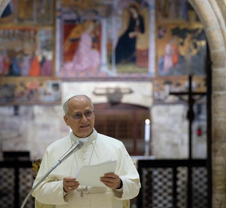 Un sacerdote in abito liturgico parla al microfono in una chiesa con affreschi sullo sfondo.