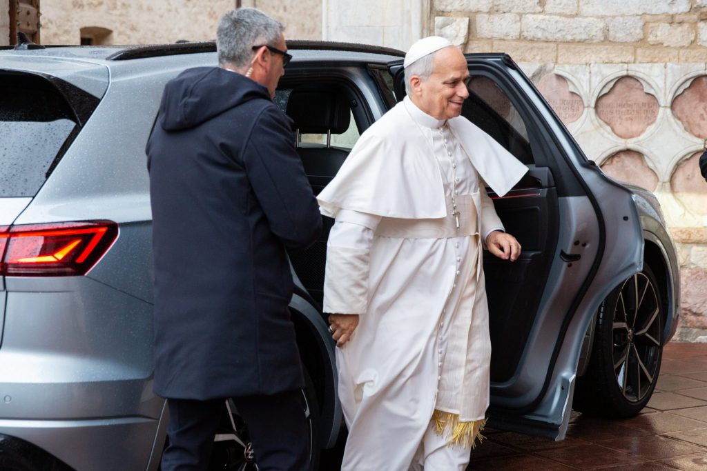 Papa Leone XIV esce da un'auto di colore grigio con un accompagnatore, sorridendo mentre si avvicina a un luogo di celebrazione.