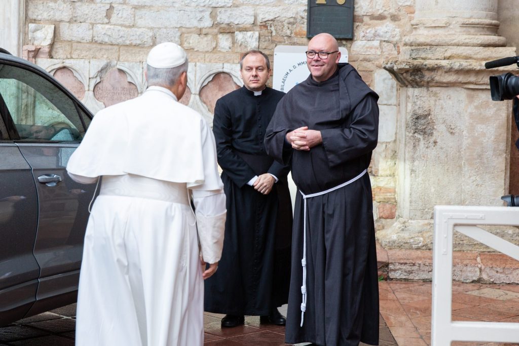 Il Papa Leone XIV in abiti bianchi, accolto da un frate in abito scuro all'esterno di un edificio, con un'altra persona in abiti scuri presente nella scena.