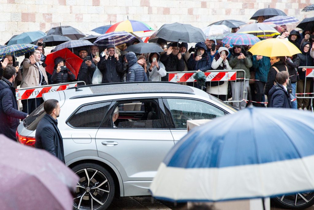 Il Papa Leone XIV arriva a Assisi, accolto da una folla di fedeli che si ripara dalla pioggia con ombrelli, mentre saluta con entusiasmo.