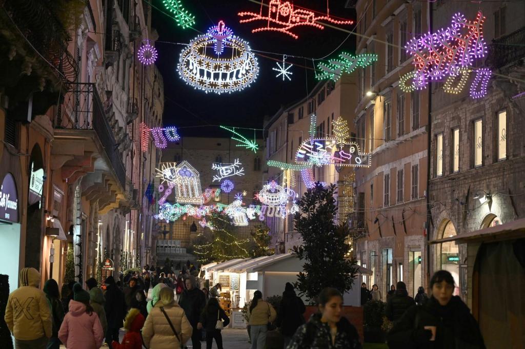 La via del centro storico di Perugia decorata con luminarie natalizie colorate, mentre un gruppo di persone passeggia. Le luci brillano tra gli edifici storici, creando un'atmosfera festosa.