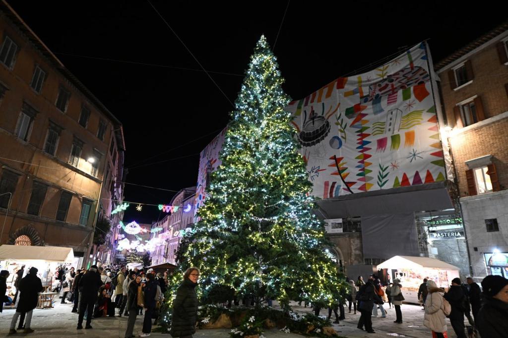 Una grande albero di Natale illuminato al centro di una piazza affollata a Perugia, con persone che passeggiano e uno sfondo di edifici storici decorati.
