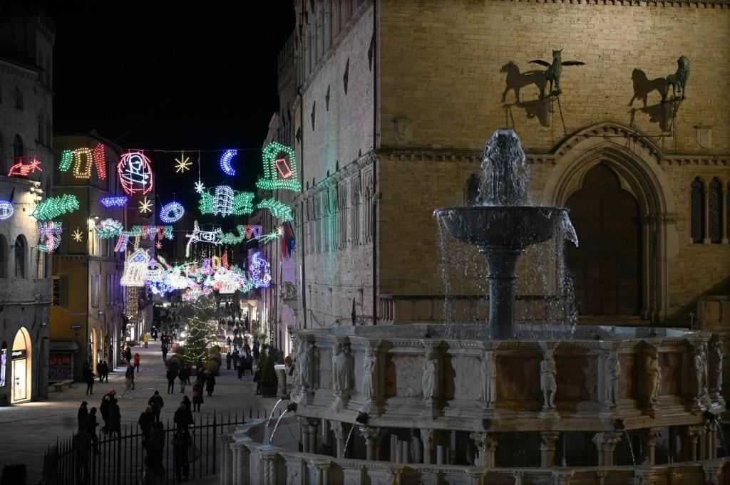 Una vista notturna di Corso Vannucci a Perugia, adornata da luminarie natalizie colorate e un albero di Natale, con una fontana in primo piano e persone che passeggiano.