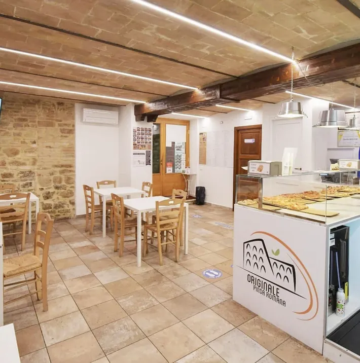 Interno della Pinseria Torre degli Sciri a Perugia, mostrando tavoli bianchi e sedie in legno, con una vetrina di pinse in primo piano.