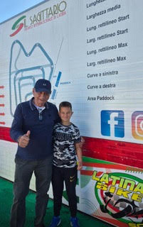 Un uomo e un ragazzo sorridono davanti a un muro informativo del circuito di karting, con segni che descrivono le caratteristiche del tracciato.