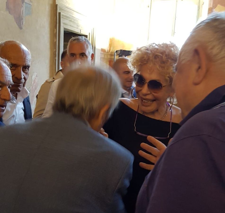 Ornella Vanoni interagisce con un gruppo di persone, sorridente e con occhiali da sole, in un ambiente interno illuminato.