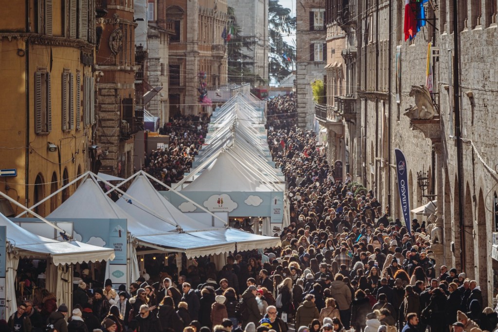 A bustling street market filled with large crowds of people walking between tents and stalls, set against historic buildings in a city atmosphere.