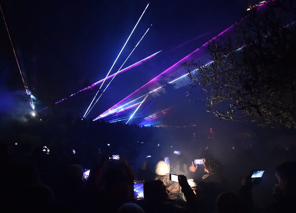 Spettacolo di luci laser durante un evento a Castiglione del Lago, con un pubblico che utilizza i propri telefoni per filmare e fotografare.