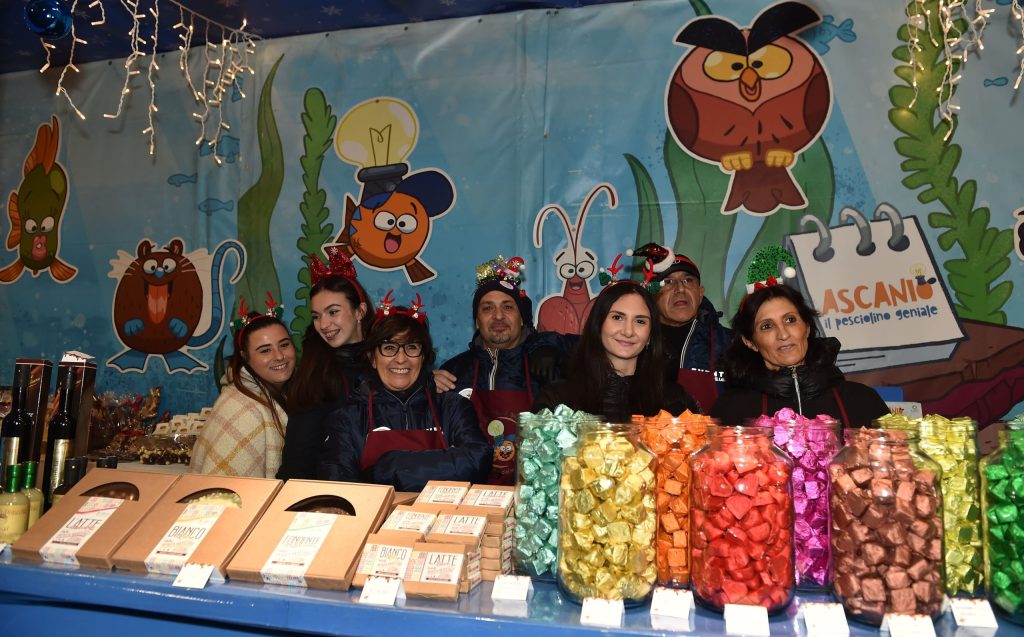 Un gruppo di persone sorridenti in un mercato natalizio, con decorazioni festive e barattoli colorati di dolciumi sul tavolo.