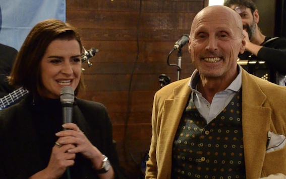 Due relatori sorridenti al microfono durante un evento, con un'atmosfera accogliente e strumenti musicali sullo sfondo.