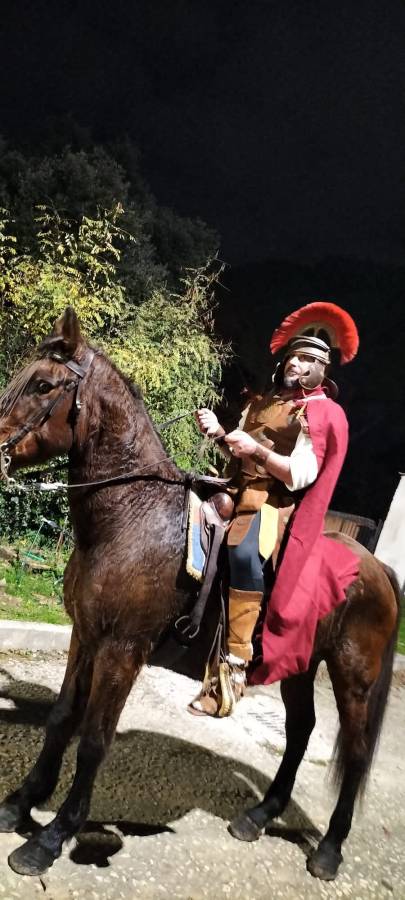Un uomo vestito da soldato romano, con elmo e mantello, monta un cavallo davanti a un ambiente scuro e illuminato da luci naturali.