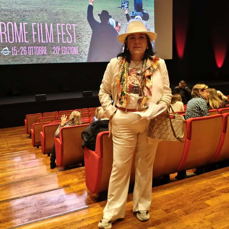 Una donna in abbigliamento elegante, con un cappello, posa in un cinema durante il Festival del Cinema di Roma, con un poster dell'evento sullo sfondo.