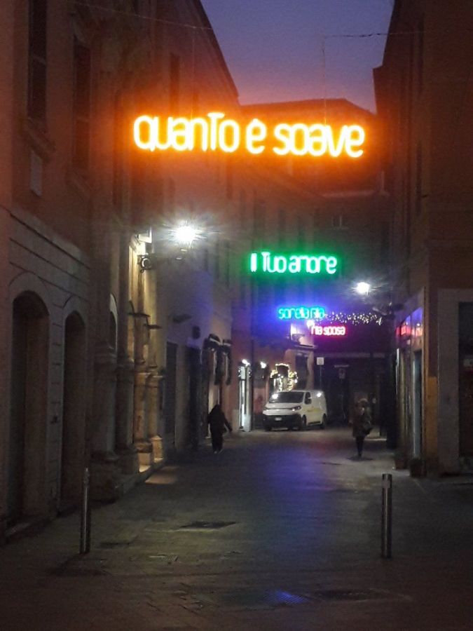 A narrow street illuminated by neon signs with the texts 'quanto e soave' in orange and 'Il Tuo amore' in green, during the evening.