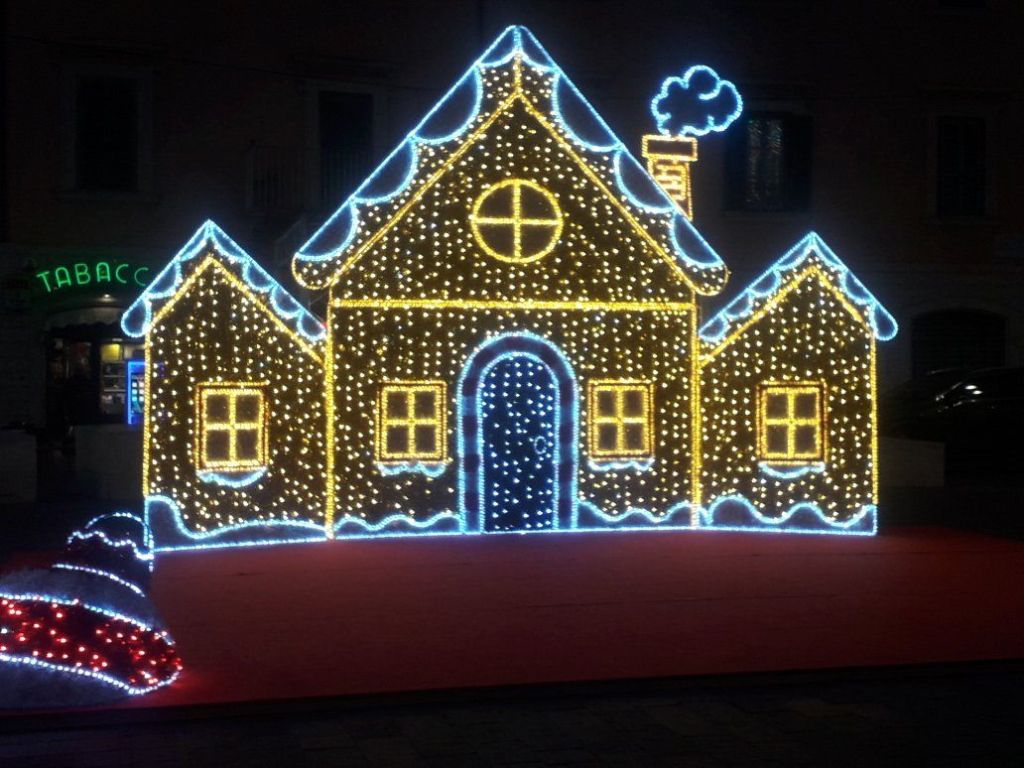 A brightly lit gingerbread house made of colorful Christmas lights, featuring a chimney and snow-like decorations on the roof, set against a dark background.
