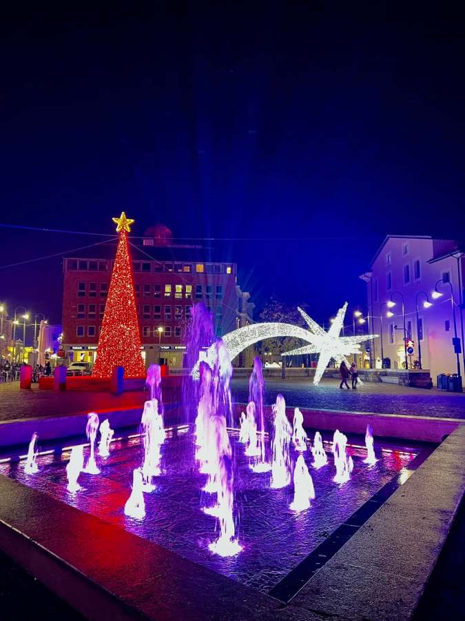 A vibrant Christmas scene at night featuring a large decorated tree with a star on top, illuminated fountains with purple lights, and a sparkling star-shaped display in a square.
