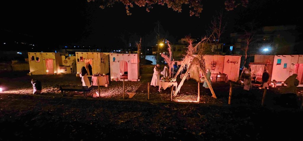 A nighttime scene of a recreated village with several illuminated tents, people dressed in traditional attire engaging in activities, and warm lighting contributing to a festive atmosphere.