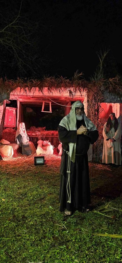 A man wearing traditional Middle Eastern clothing stands in front of a dimly lit nativity scene, with a small group of other figures in the background. The scene is illuminated with a warm, red light.