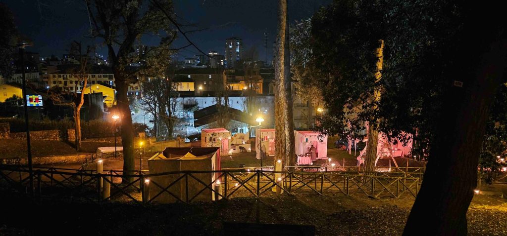 Nighttime view of a park with lit tents and a city skyline in the background.