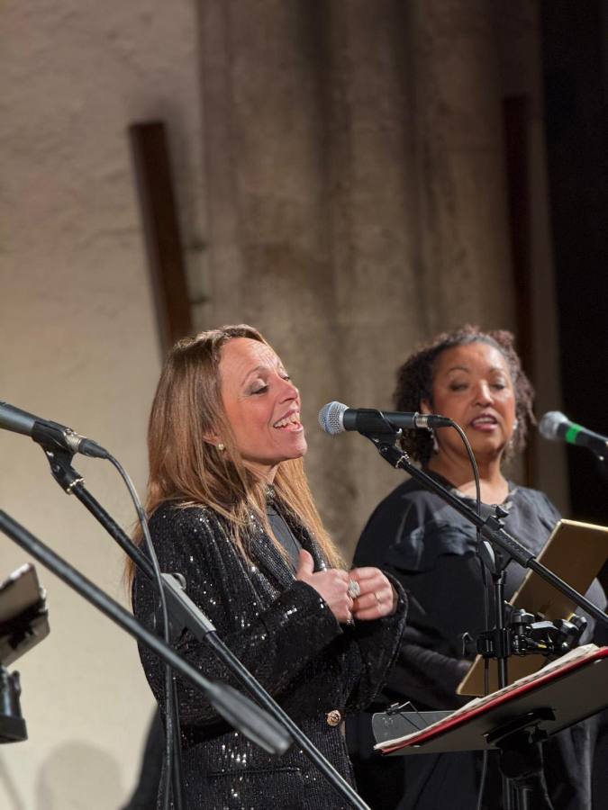 Due cantanti sul palco durante un concerto, una di fronte all'altra, mentre cantano con microfoni in mano. L'ambientazione è elegante e suggestiva.