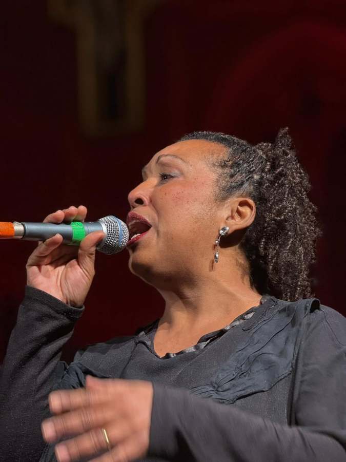 Cantante al concerto di Mario Donatone & Bluesman Latino, con microfono in mano mentre interpreta una canzone, sullo sfondo una chiesa illuminata.